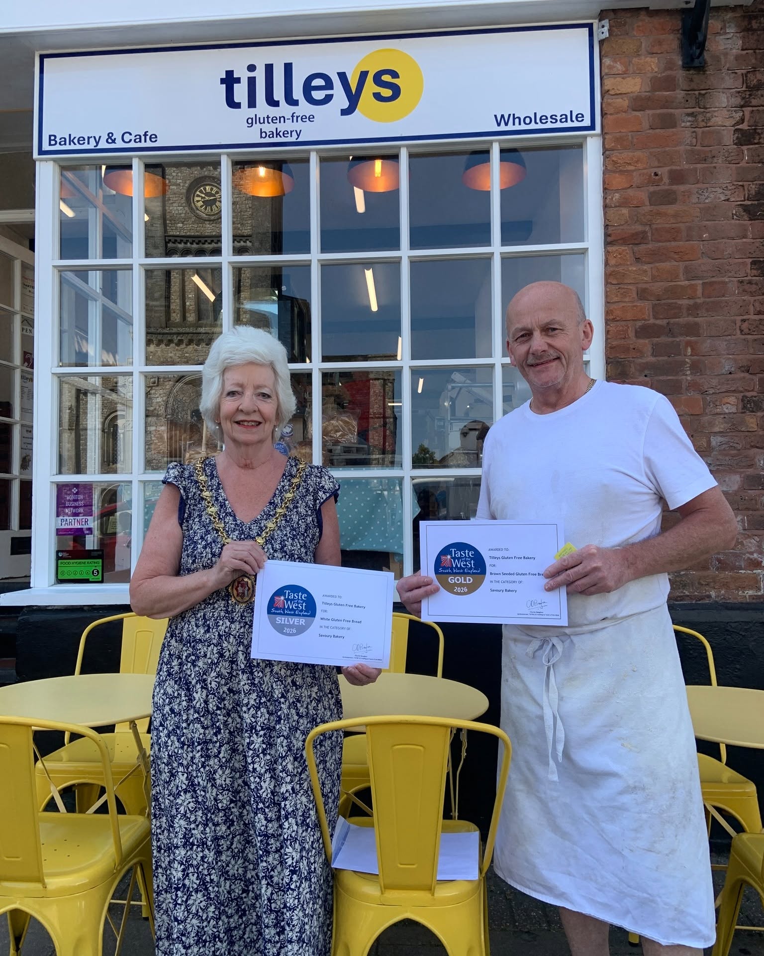 Brian Spence with the Mayor of Honiton holding Taste of the West Gold and Silver 2026 certificates outside Tilley's Gluten Free Bakery